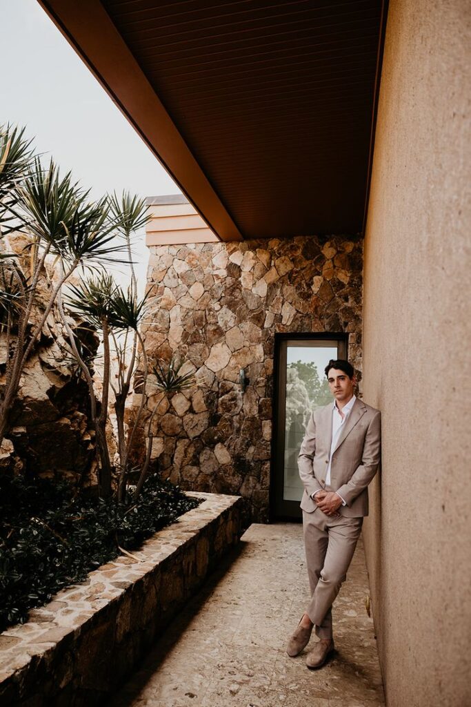 Groom in a tan suit poses, leaned against a wall before his wedding.